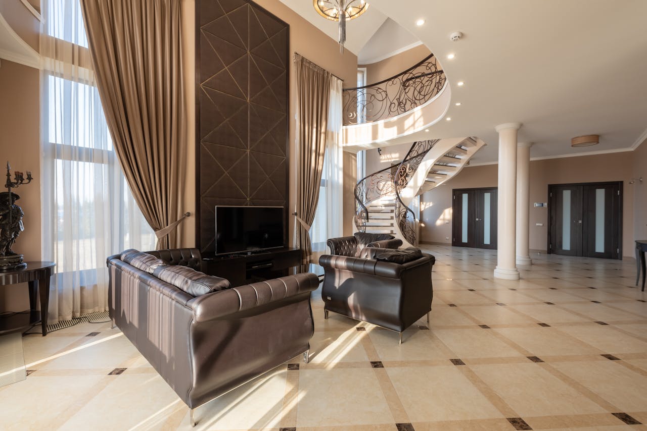 Soft leather black couches on tiled floor near window with curtains in modern stylish house with stairway and doors near columns